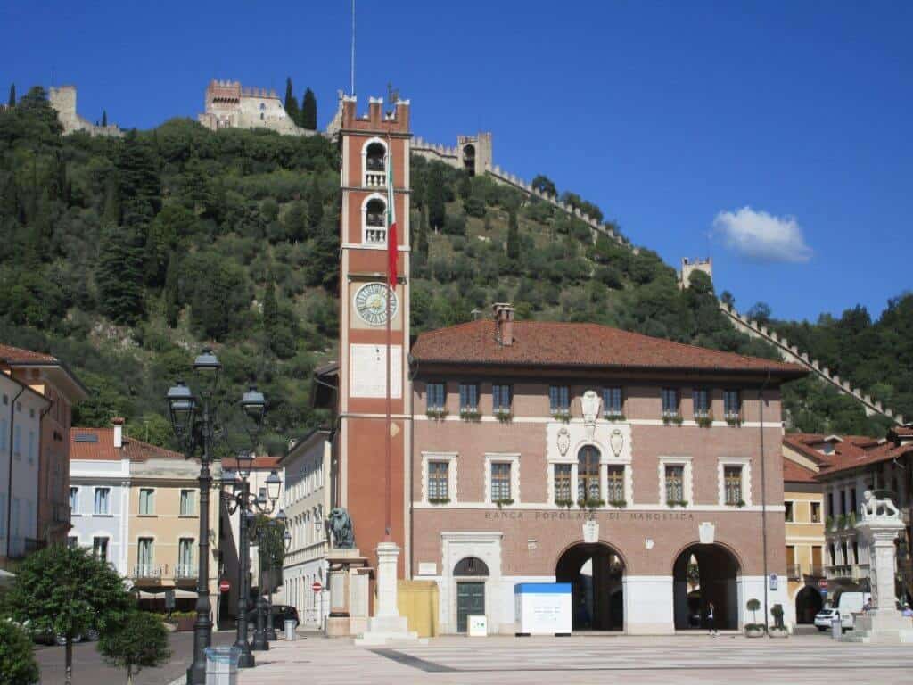 The charming town hall of Malcesine, Italy, with its distinctive red and white clock tower, stands prominently in the main square. Lush green hills and the historical Scaligero Castle loom in the background under a clear blue sky, exemplifying the scenic beauty of this lakeside town on Lake Garda.