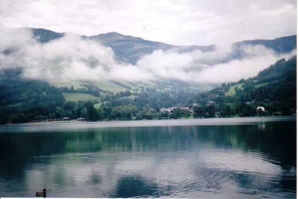 Tranquil view of Zell am See, Austria, featuring the glassy surface of the lake reflecting low clouds nestled in the surrounding mountains. The lush green slopes of the mountains descend into the calm waters, while a small part of the village is visible across the lake, adding a quaint charm to the scenic landscape.