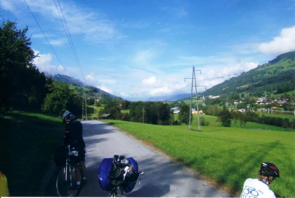 Cyclists navigating the Tauern Cycle Path near Zell am See, Austria, with scenic views of lush green fields and rolling hills under a partly cloudy sky. The trail runs alongside a country road, framed by utility poles, showcasing the rural beauty of the Austrian countryside. In the background, small houses and a village nestle against a backdrop of forested mountains