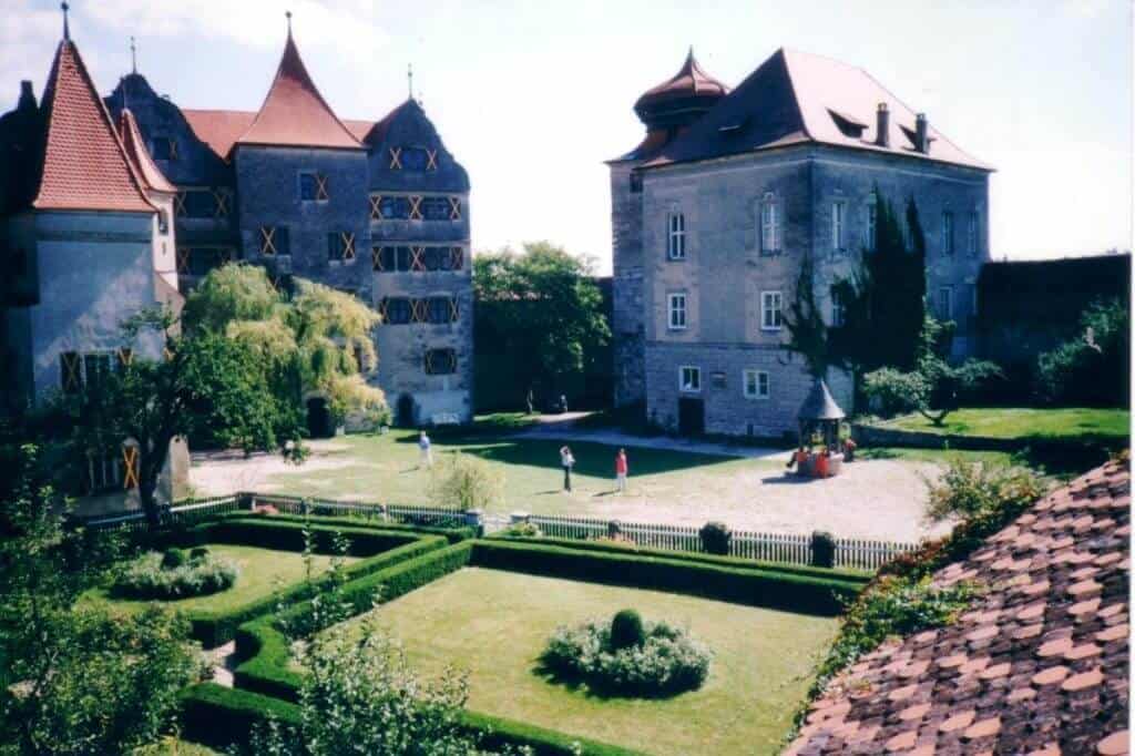 A captivating view of Harburg Castle in Germany, featuring its majestic stone architecture with distinctive half-timbered patterns. The castle is surrounded by manicured gardens and a well-maintained lawn, where visitors are seen enjoying the serene environment.