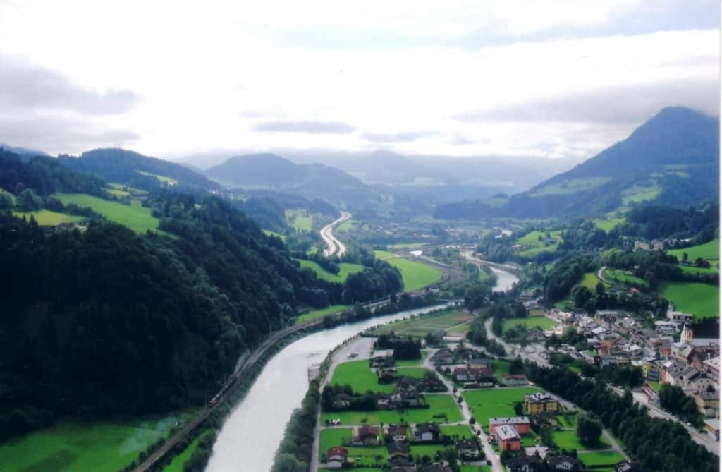 Aerial view of Werfen, Austria, showcasing a winding river that cuts through lush green valleys and a small picturesque town. The river is bordered by dense forests and agricultural fields, with mountains rising dramatically in the background. The cloudy sky casts soft shadows over the landscape