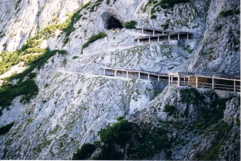Rugged pathway and wooden walkway leading to the entrance of the Eisriesenwelt Cave in Austria. The path carves through a steep, rocky mountainside, with sparse vegetation clinging to the gray limestone. This challenging route offers adventurous access to the world's largest ice cave, nestled within the imposing Alps