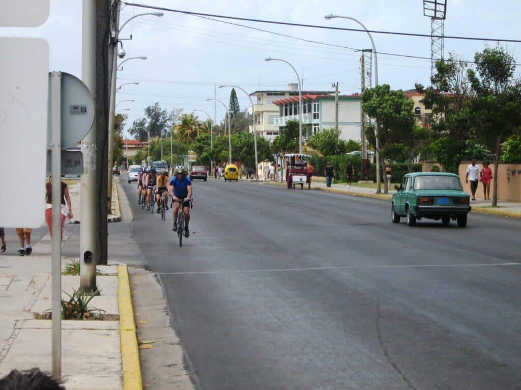 A group of cyclists in various shades of sportswear ride along First Avenue in Varadero, Cuba. They share the road with a classic green car and pedestrians on the sidewalk, set against a backdrop of residential buildings and lush greenery. The scene captures a typical sunny day in Varadero with light traffic and clear skies.