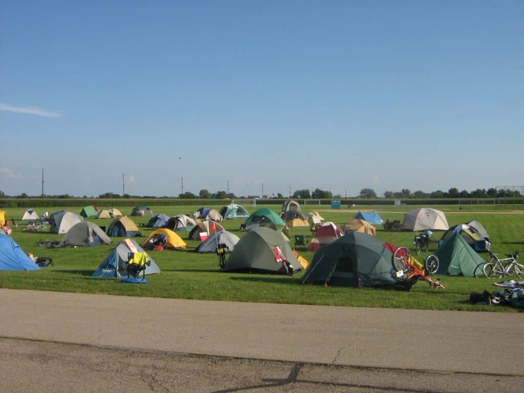 A temporary campground set up on a vast grassy field in Waupun, during one of the Wisconsin bike tour . The scene is dotted with numerous colorful tents ranging from blue, yellow, green, to gray, under a clear blue sky. Bicycles and camping gear are scattered around, indicating an active outdoor adventure. The flat, open landscape in the background emphasizes the rural setting of this camping event.