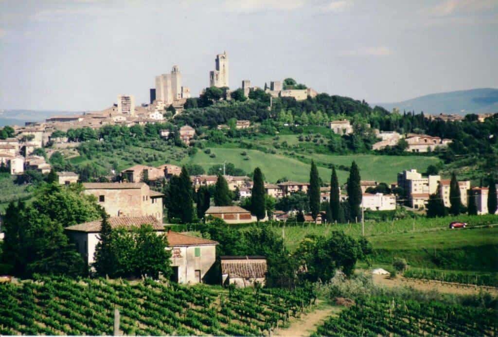 Bicycle touring in Italy will perhaps include Tuscany. A scenic view of the hilltop town of San Gimignano, Italy. The town is characterized by its medieval towers and buildings nestled among rolling green hills. In the foreground, lush vineyards and rows of cypress trees add to the picturesque rural landscape.