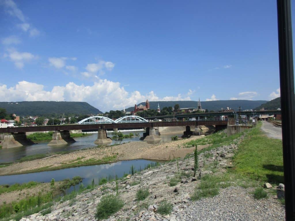 View of Cumberland, PA, showcasing a serene river with multiple bridges, including a distinctive blue arched bridge, against a backdrop of a small town and rolling hills under a blue sky with fluffy clouds.
