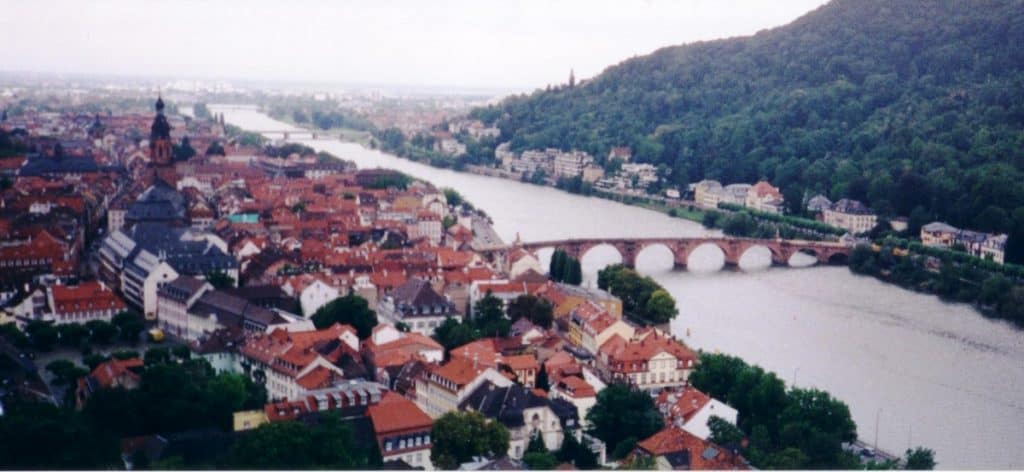 Aerial view of Heidelberg, Germany, showing the historic cityscape along the Neckar River. The image features the Old Bridge with its classic arches, prominent church spires, and a mix of red-roofed buildings set against a backdrop of lush green hills. This picturesque setting captures the essence of a traditional European city nestled in a river valley.