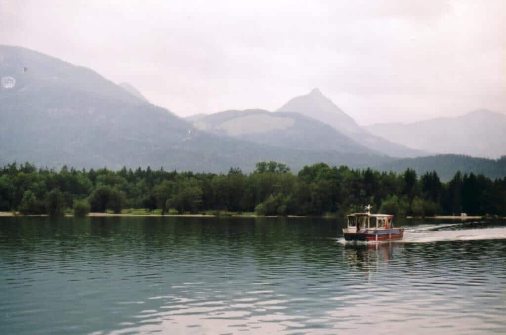 The image features a tranquil lake setting with a small ferry boat cruising across calm waters towards a lush, tree-lined shore. In the background, majestic mountains partially shrouded in mist rise dramatically, enhancing the serene and picturesque landscape. The overcast sky adds a moody atmosphere to the peaceful scene, typical of a lakeside setting in a mountainous region.