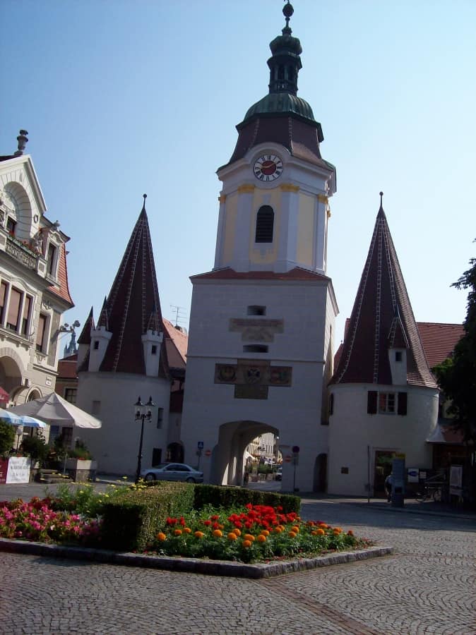 The historical entrance gate to the walled town of Krems, Austria, featuring a clock tower and flanked by pointed rooftops, with a vibrant flower bed in the foreground, paving the way to the old town streets