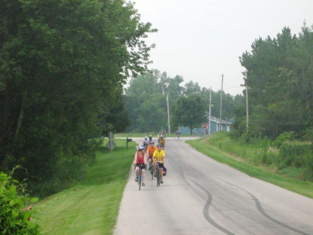 A group of cyclists in colorful attire rides along a quiet country road near Waupun, Wisconsin. The road, flanked by lush greenery and mature trees, curves gently into the distance, where more cyclists can be seen.