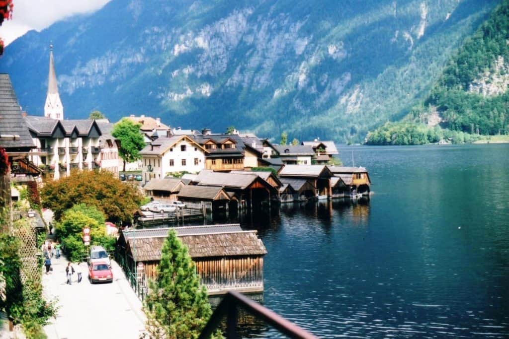 The image captures the charming lakeside view of Hallstatt, Austria. Colorful traditional houses with alpine architectural details line the lush, tree-covered bank of the lake, with a prominent church spire rising against the backdrop of steep, forested mountains. Wooden boathouses stretch out over the clear blue waters, contributing to the picturesque and tranquil atmosphere of this quaint village.