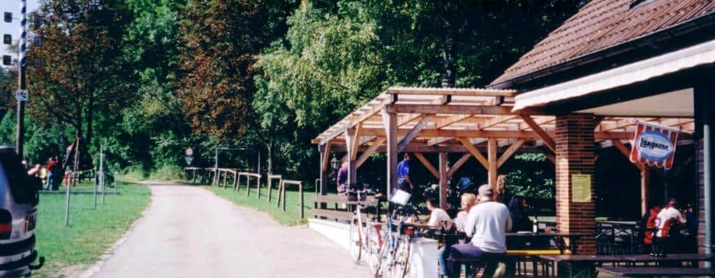 Cyclists and visitors enjoy a sunny day at a roadside cafรฉ near Augsburg, Germany, located along the Romantic Road bicycle path. The cafรฉ features an outdoor seating area under a wooden pergola, surrounded by lush greenery and a relaxed atmosphere.