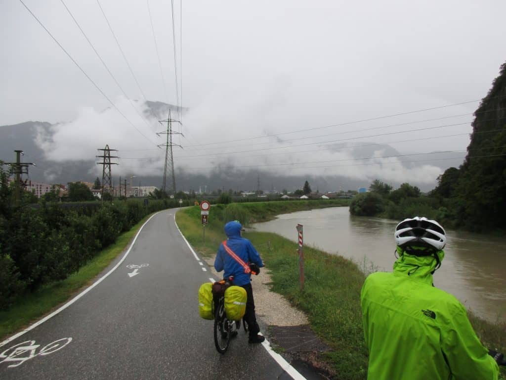 This photo captures a moment on the Adige Cycling Path near Bolzano, Italy, under overcast skies. Two cyclists, one in a blue jacket and one in a bright green jacket, ride along a well-maintained bike lane beside a river. The landscape features lush greenery on either side of the river, with mist-covered mountains in the background and power lines running parallel to the path