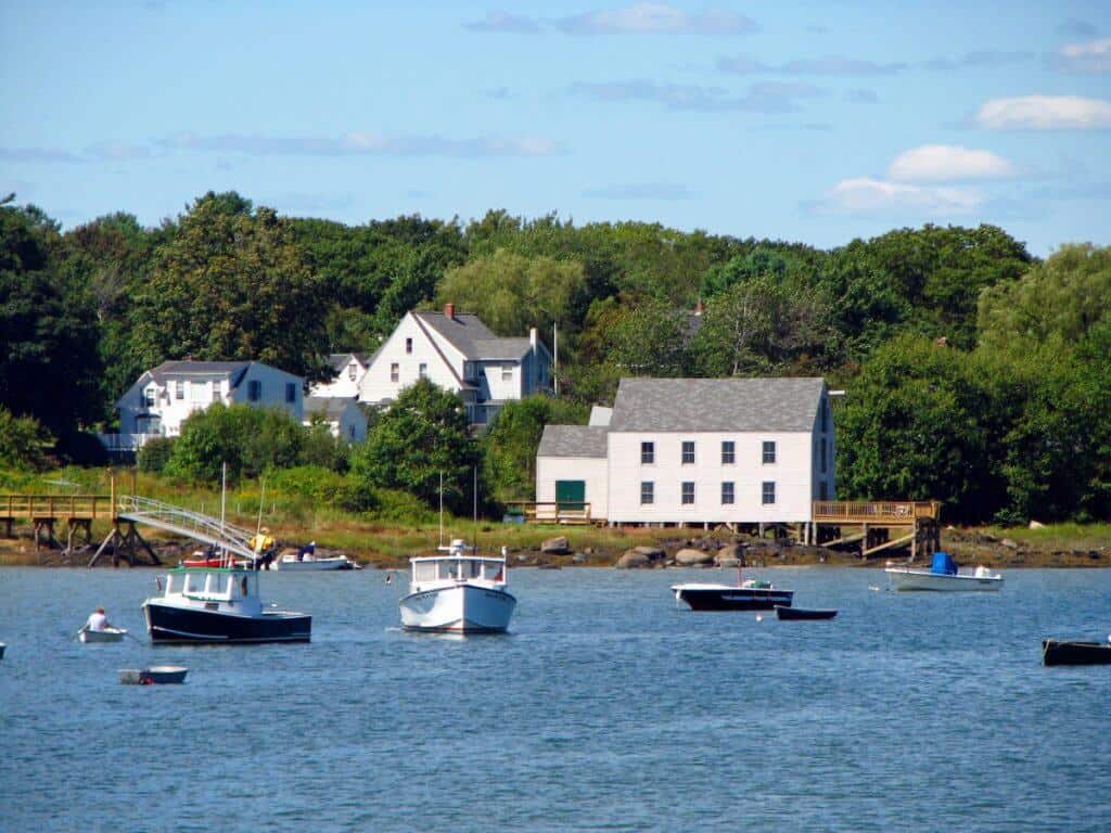 Quaint waterside view of Cape Porpoise, Maine, featuring small boats bobbing in the calm waters, traditional New England white houses, and lush trees in the background, reflecting a tranquil coastal village lifestyle.