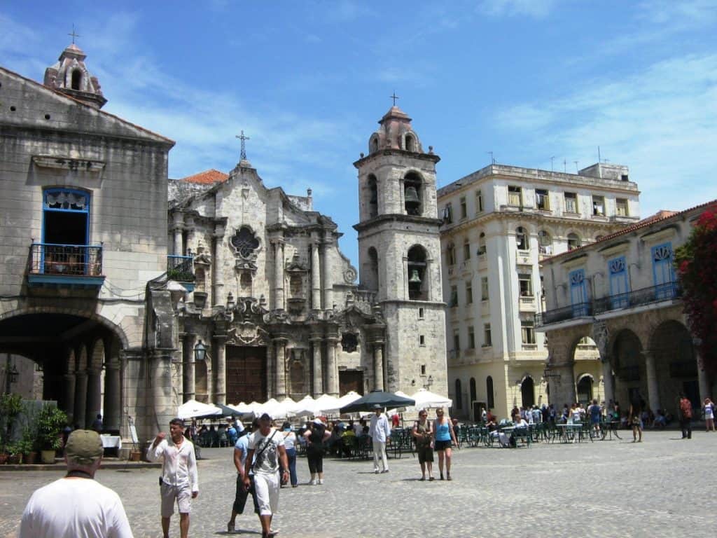 Busy square in front of the Havana Cathedral in Cuba, featuring the cathedral's intricate façade and distinctive bell towers. The square is bustling with tourists and locals, with several sidewalk cafes under sun umbrellas. Historical buildings with vibrant blue balconies surround the cathedral, under a clear blue sky.