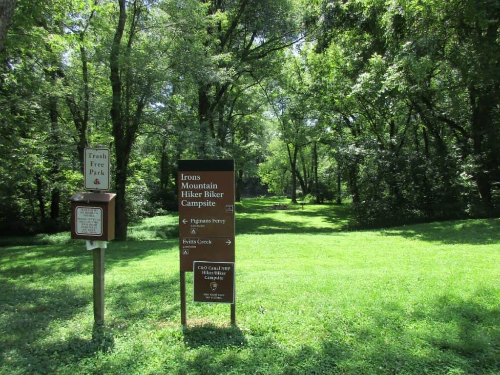 The Irons Mountain Hiker Biker Campsite signpost on the C & O Trail, indicating a trash-free park, with a backdrop of verdant trees and a well-maintained grassy area with a picnic table in the distance.