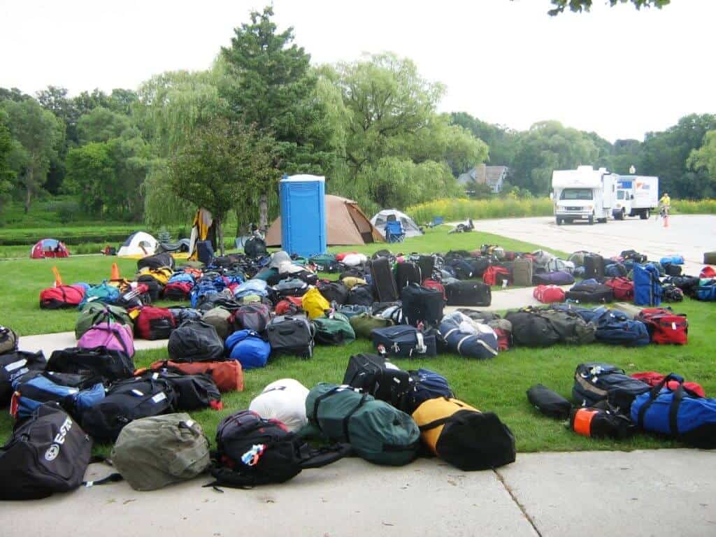 A large collection of assorted luggage, including backpacks and duffel bags, neatly arranged on a grassy area during a bike tour in Wisconsin. The bags come in various colors and sizes, indicating the diverse group of participants. In the background, tents and a portable restroom can be seen, with a medical service vehicle parked nearby, emphasizing the organized and well-supported nature of the tour.