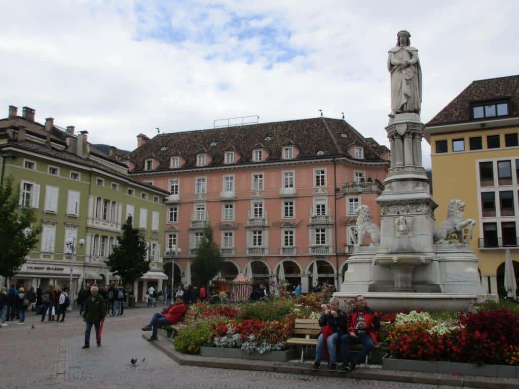 This image shows the bustling main square in Bolzano, Italy, featuring a mix of historical and vibrant architecture. In the center stands a statue of a robed figure on a pedestal, surrounded by colorful flower beds and flanked by stone lions. The square is lively with people walking and others sitting on benches, enjoying the scene. Prominent buildings, including the pastel-hued Stadt Hotel, frame the square, enhancing the European charm of the setting.
