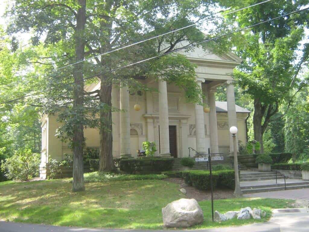 Historic building at the Chautauqua Institute, NY, surrounded by lush greenery and shaded by tall trees. This classic structure features a large portico with four imposing columns and a pediment, enhancing its grand neoclassical design. The building is set back from a quiet street, framed by power lines overhead and large rocks and a street lamp at the foreground.