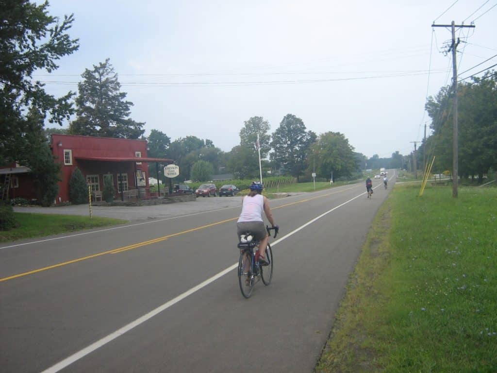 Cyclists riding along a rural road in America's Grape Country, New York State. The road is lined with utility poles and stretches past a red building labeled 'Hometown Market' on a hazy day. The landscape is verdant with trees and grass along the roadside