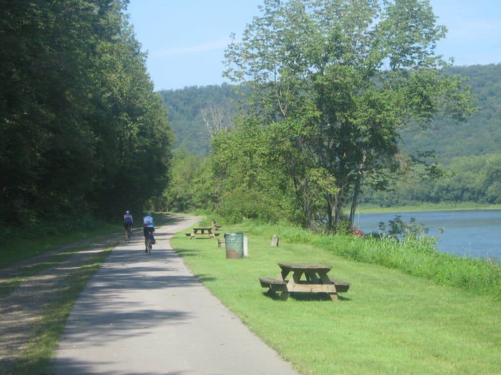 Cyclists enjoying a ride on the scenic Allegheny River Trail in Pennsylvania. The trail is flanked by dense green trees on one side and the calm Allegheny River on the other, under a clear blue sky. Picnic tables and trash bins along the path