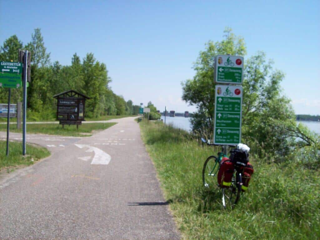 A touring bicycle loaded with red panniers stands beside directional signage on the Danube cycle path near Ottensheim, Austria, with the serene river and lush greenery in the background on a sunny day.