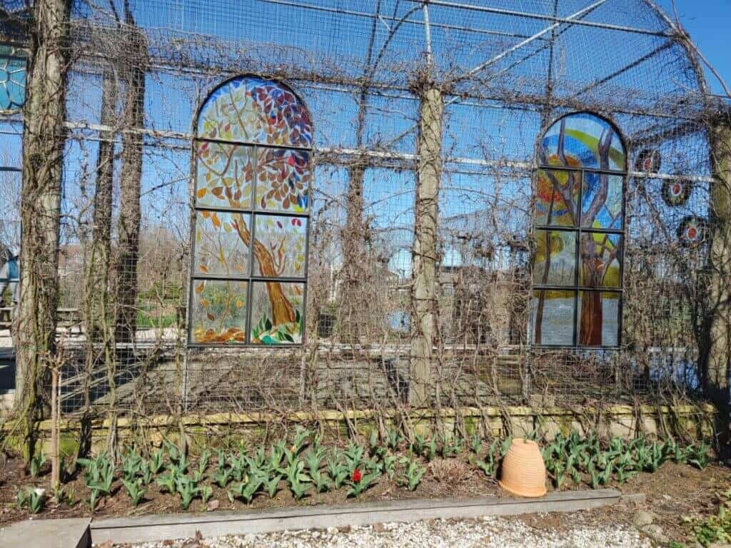 An outdoor art exhibit along a Dutch canal, featuring two large stained glass panels depicting vibrant trees and landscapes. The panels are set within a bare trellis structure, with emerging tulip plants at their base, blending art with the budding signs of spring.