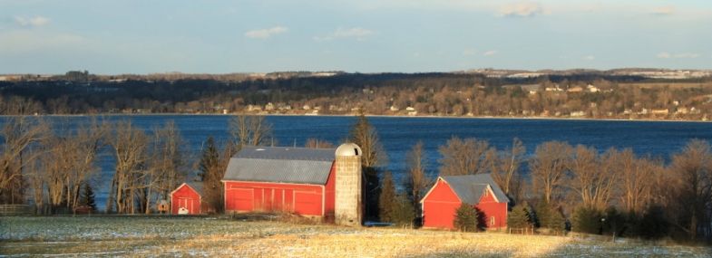 A picturesque winter scene overlooking Owasco Lake in the Finger Lakes region, featuring a prominent red barn and silo surrounded by smaller buildings, all set against a backdrop of the lake. The trees, bare from the cold season, and a light dusting of snow on the ground accentuate the vibrant red of the farm structures under a clear sky