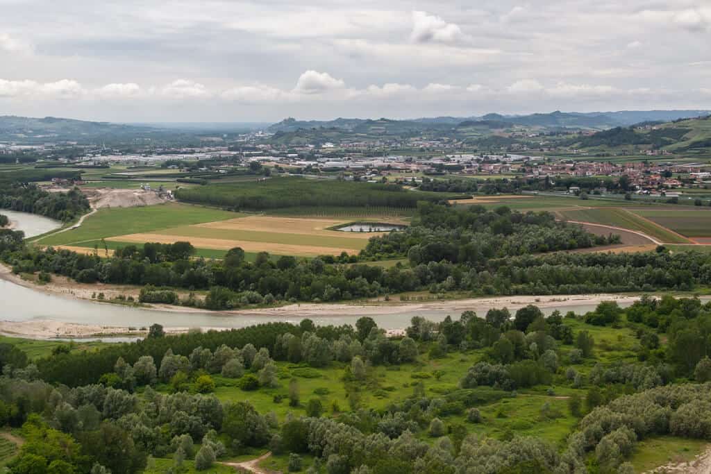 An expansive view of the Piedmont countryside in Italy, featuring a winding river flanked by lush greenery and agricultural fields. The landscape includes patches of forests, fields in various shades of green and brown, and distant towns.