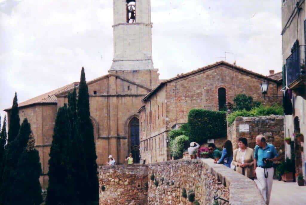 Visitors walking by the stone walls of a church in Pienza, Tuscany, with its distinctive bell tower reaching into a cloudy sky. The ancient architecture of the church and surrounding buildings are typical of the historic charm found in this region.