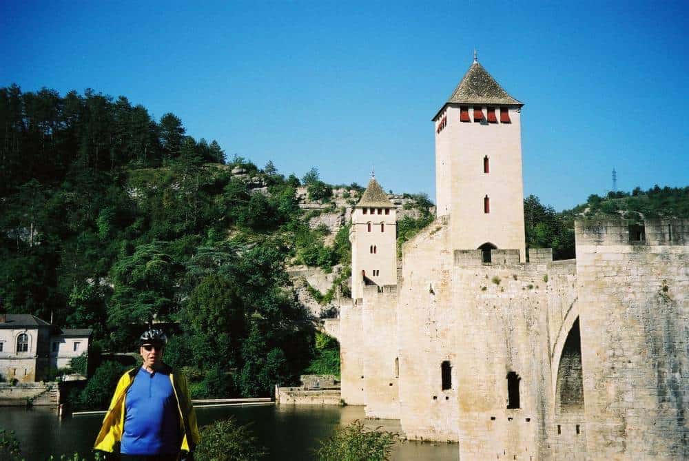 A person in cycling gear stands in front of the historic Pont Valentrรฉ, a fortified stone arch bridge crossing the Lot River in Cahors, France, with its prominent towers under a clear blue sky