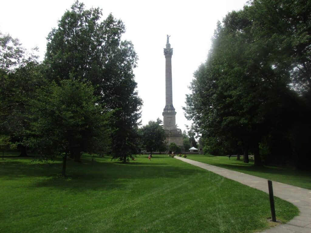 Brock Monument at Queenston Heights, Ontario, standing tall amidst a lush green park. The historical column, crowned with a statue, dominates the landscape, serving as a memorial to Major General Sir Isaac Brock. A clear pathway leads to the monument, inviting visitors to approach and reflect on the historical significance of the site. The surrounding greenery and towering trees enhance the serene and dignified atmosphere of this important Canadian landmark.