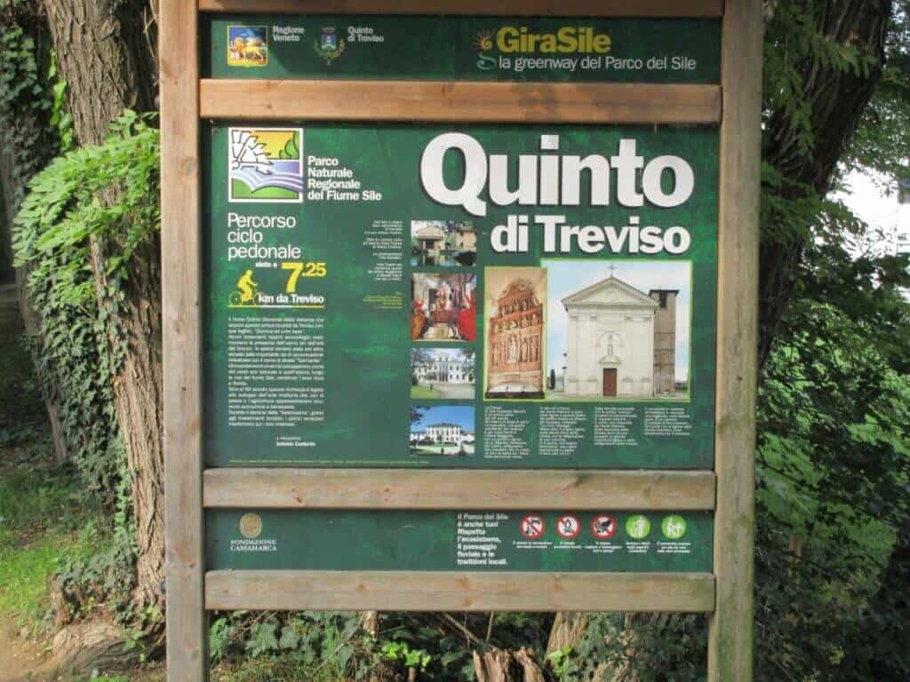 An informative signboard for the Quinto di Treviso cycling route, displaying maps, photographs of local attractions, and route details for the GiraSile greenway, nestled among lush greenery.