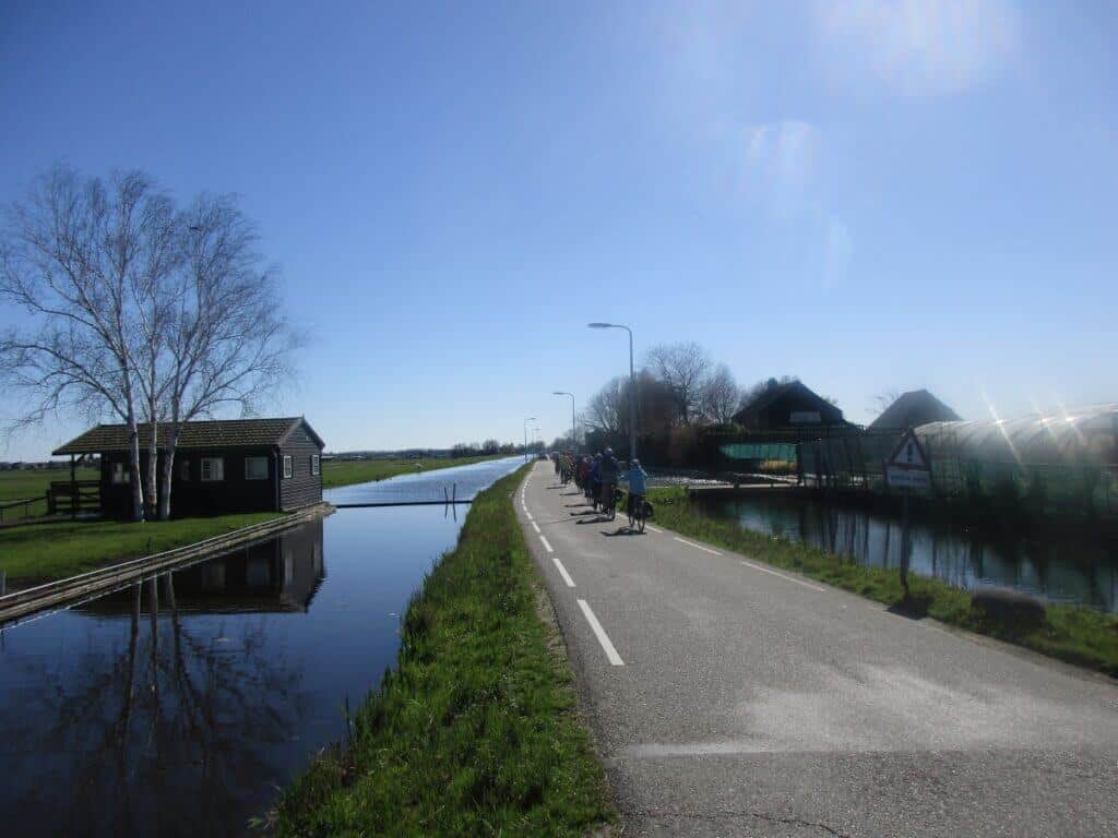 A group of cyclists embarks on a sunny ride along a dike in the Netherlands, with clear reflections on the calm canal water and a traditional Dutch house nestled among leafless trees.