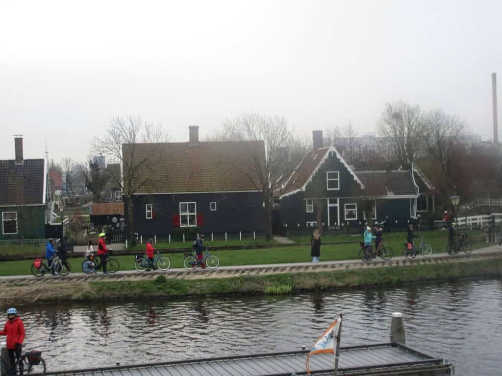 Cyclists begin their journey departing from the quaint village of Zaanse Schans, riding along a canal with traditional Dutch houses providing a picturesque backdrop. The overcast sky and calm water reflect the serene ambiance of this historic region in the Netherlands.