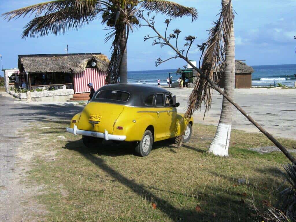A vintage yellow car parked on a coastal road near Havana, Cuba, with a rustic beach bar and palm trees in the background. The ocean is visible in the distance, and a few people can be seen enjoying the sunny beach day. The scene captures a relaxed, tropical atmosphere typical of Cuban coastal locales.