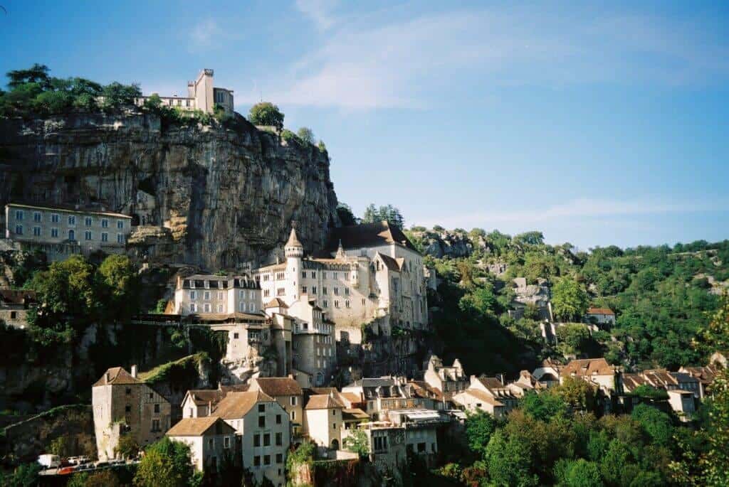 The ancient village of Rocamadour in France, built into the side of a cliff, with historic multi-storied buildings rising steeply above the Alzou canyon, under a clear blue sky.