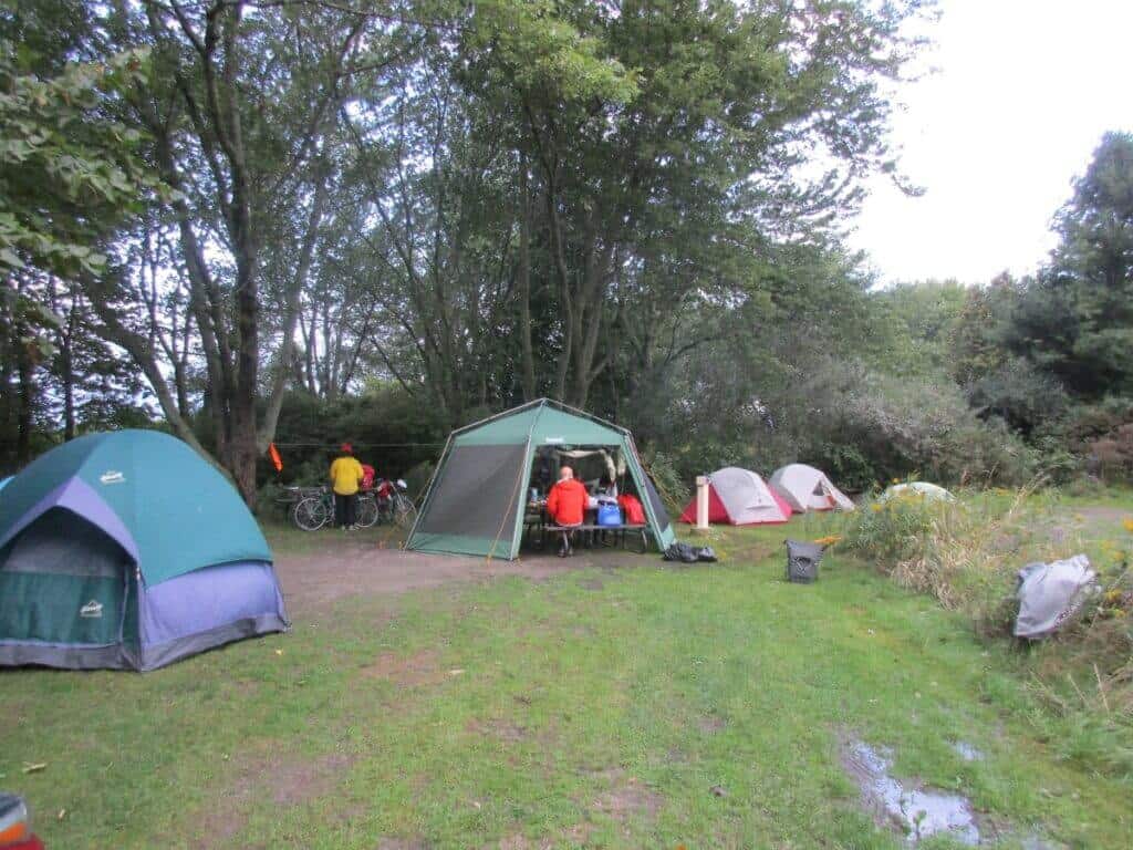 Camping scene at Rock Point Provincial Park in Ontario. Multiple tents are set up on a grassy area surrounded by dense trees, creating a cozy and secluded camping environment. A central kitchen tent serves as a communal gathering spot where campers are seen engaging in meal preparations and conversations. Bicycles equipped with touring gear are parked nearby