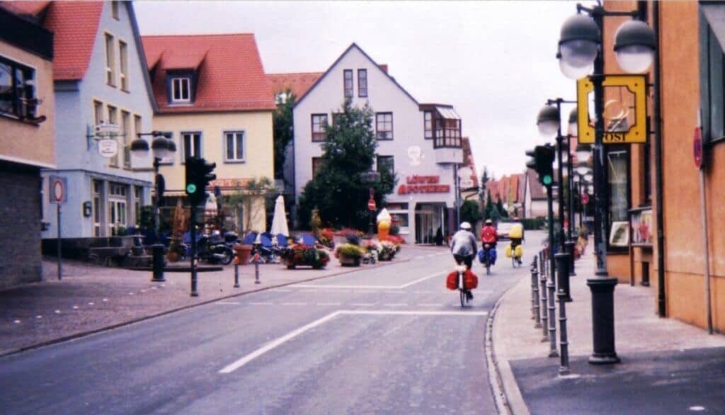 Cyclists touring a quaint town along the Romantic Road Bicycle Touring Route in Germany. The street scene captures a lineup of charming, colorful buildings, street lamps, and a clear road marked with cycling lanes. The cyclists, equipped with gear and brightly colored clothing, add a lively touch to the peaceful town setting.
