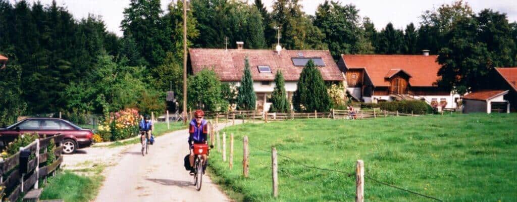 Cyclists journey along the Romantic Road Bicycle Route in the Bavarian countryside, framed by lush meadows and dense forests. A picturesque row of rural houses lines the path, complete with blooming gardens and well-kept yards.