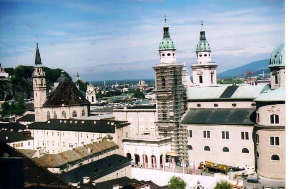 The image offers a panoramic view of a prominent cathedral complex in Salzburg. The photograph captures multiple historic buildings with intricate architectural details, including spired towers and domed rooftops. One of the cathedral's towers is partially scaffolded, indicating restoration work. The surrounding area includes several other buildings with similar architectural styles, set against a backdrop of the city extending into distant mountains.