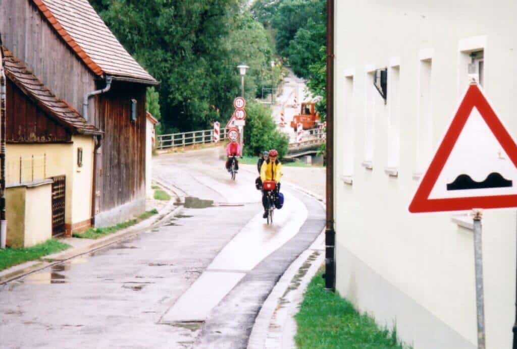 Cyclists navigating a wet road through the village of Schillingsfรผrst, part of the Romantic Road Cycle Route in Germany. The scene captures them riding past traditional houses and under traffic signs, with lush greenery in the background. This image showcases the blend of adventure and quaint rural charm that the route offers.