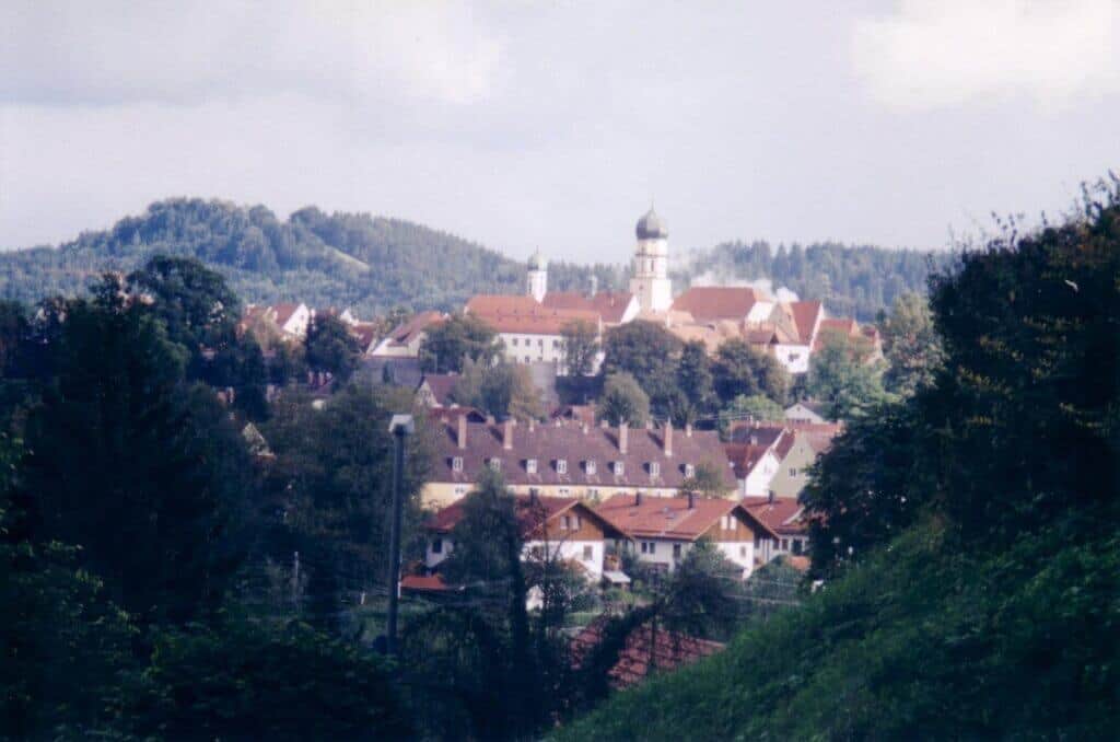 A scenic overlook of Schongau, Germany, nestled among lush, rolling hills. The town features clustered red-roofed houses and a prominent white church tower rising above the verdant landscape.
