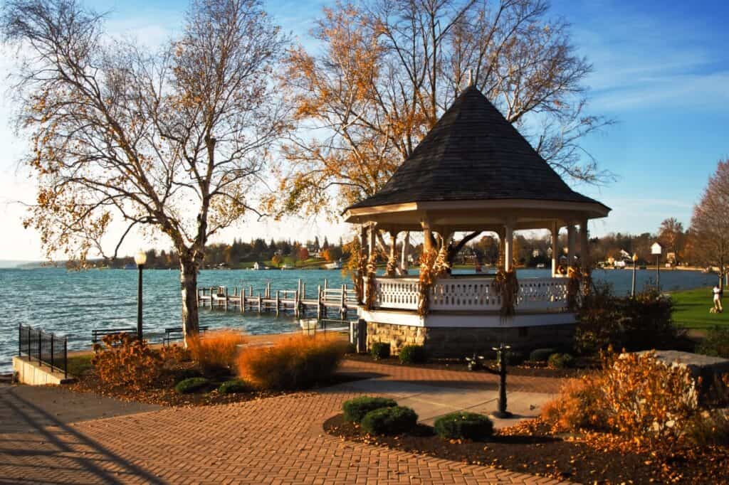 A serene autumnal scene at a small park on the shores of Skaneateles Lake, showcasing a charming gazebo adorned with seasonal decorations. The gazebo, surrounded by a brick pathway and ornamental grasses turning golden in the fall, overlooks the clear blue waters of the lake