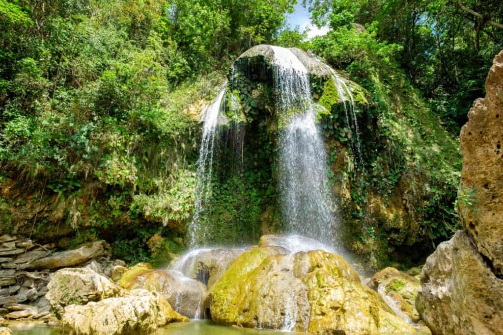 Picturesque view of a cascading waterfall at Soroa, Cuba. The waterfall spills over lush, moss-covered rocks into a serene pool below, surrounded by dense tropical foliage. Sunlight filters through the canopy, highlighting the vivid greenery and the shimmering water.