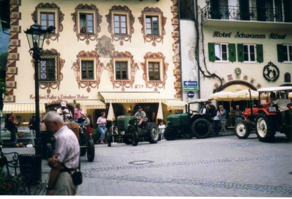 The image depicts a bustling street scene in St. Wolfgang with ornate, traditional buildings. The façade of the building on the left is adorned with decorative window frames and frescoes, and it's labeled "Konditorei Kaffee & Schokoladen Manufaktur." Adjacent is the "Hotel Schwarzes Rössl," characterized by its green shutters and a quaint balcony. The street is lively with people and a lineup of vintage tractors, suggesting a local festival or event.