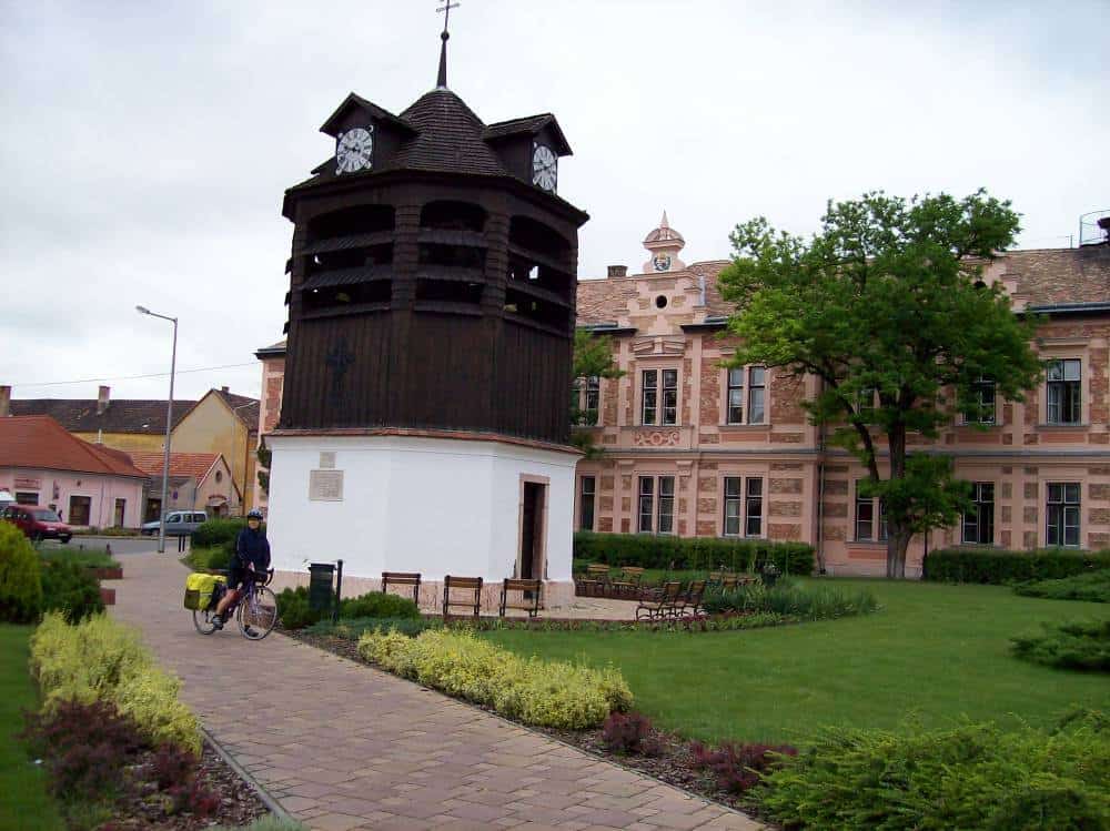 A cyclist rides past the historic Haranlab wooden clock tower in Tata, Hungary. The tower stands prominently in a well-manicured public square with patches of green grass and landscaped bushes, adjacent to a classical building with intricate facade details