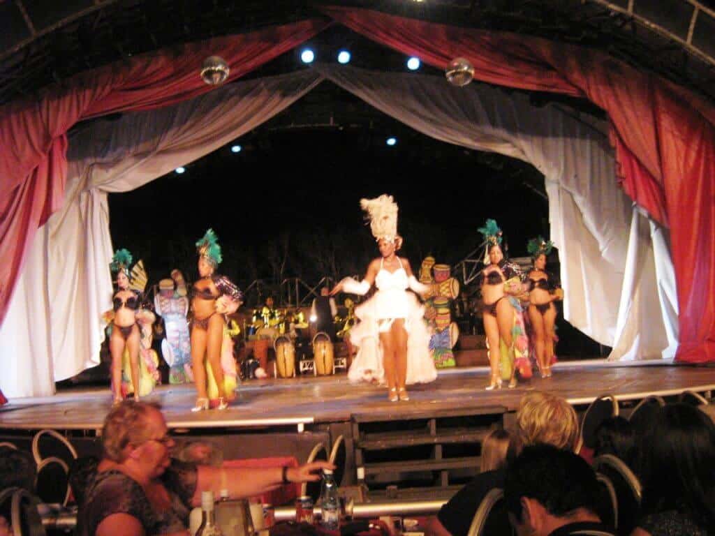 A vibrant live performance at Tropicana Varadero in Cuba, featuring dancers in colorful costumes with feathered headpieces. The stage is festively decorated with drapes and tropical accents, under atmospheric blue stage lighting. Audience members seated at tables enjoy the show, engaging with the dynamic cultural display.