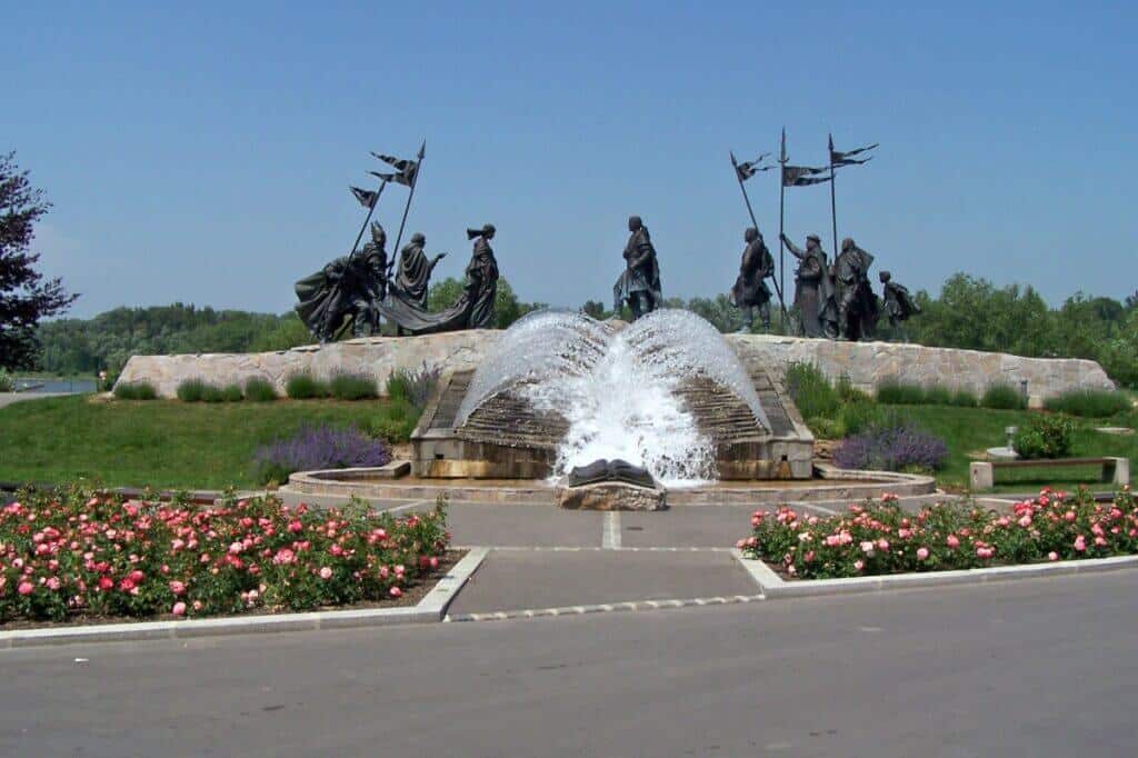 Dynamic statues in Tulln, Austria, depicting a group of figures with flags, set against a backdrop of a water fountain, surrounded by manicured flower beds and greenery, under a clear sky.