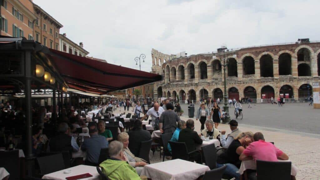 This image captures a vibrant scene from the main square in Verona, Italy, showcasing the lively atmosphere of a café-lined street. People are seated under large umbrellas at outdoor tables, enjoying meals and conversations. In the background, the historic Roman amphitheater, known for its arches and ancient stone construction, adds a grand architectural element to the bustling urban environment. The square is filled with pedestrians and cyclists